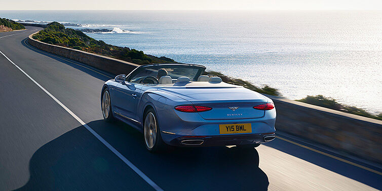 Bentley Almaty New Bentley Continental GTC Azure convertible rear view in Light Windsor blue paint with 22 inch ten spoke wheels driving dynamically on an ocean road in the sunshine