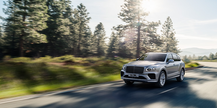 Bentley Almaty Bentley Bentayga SUV front three quarter in Moonbeam silver paint driving dynamically along a tree-lined road